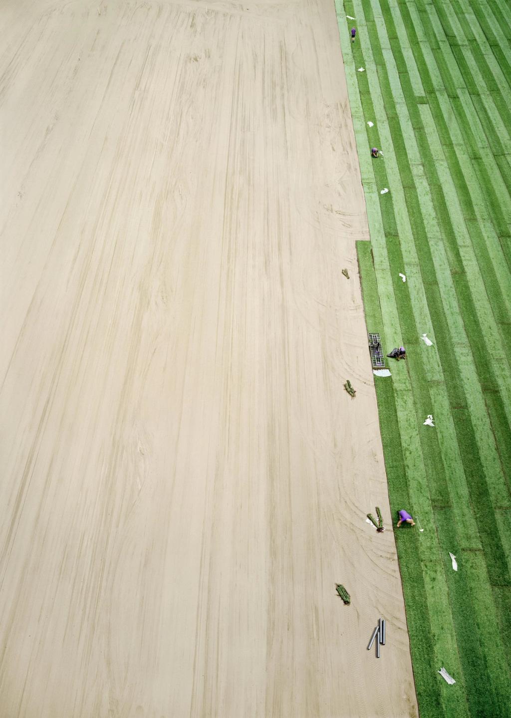 Aerial view of turf-laying: a sandy field and strips of green sod, with workers