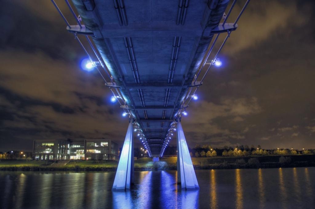 View from below of the Infinity Bridge deck at night, with blue lanterns and supports in the water