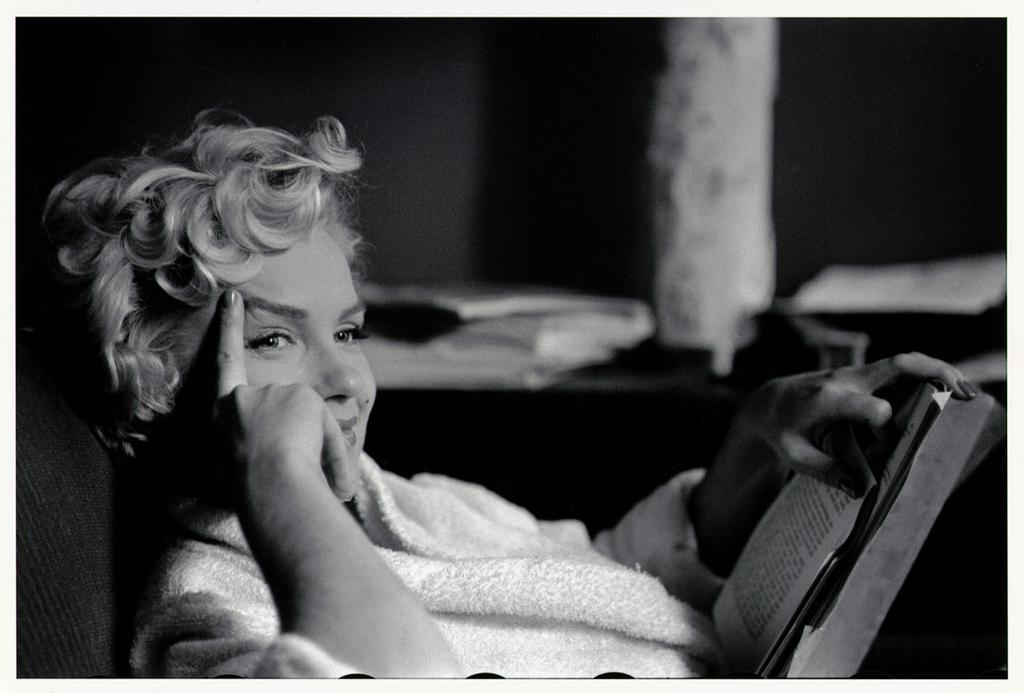 Marilyn Monroe in a light robe, smiling as she reads a book, resting her head on her hand