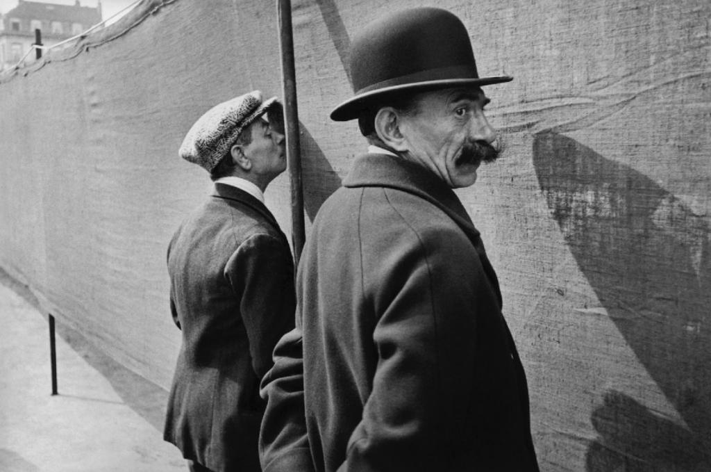 Two men by a fabric barrier; one in a bowler hat with a moustache glances back, the other in a cap leans against it