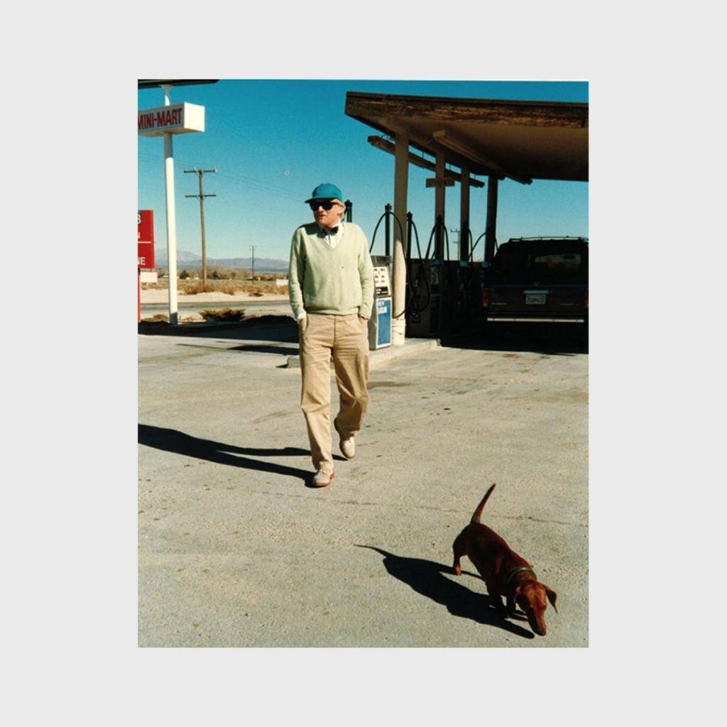 An older Hockney in a cap walking past a petrol station, a dachshund at his side