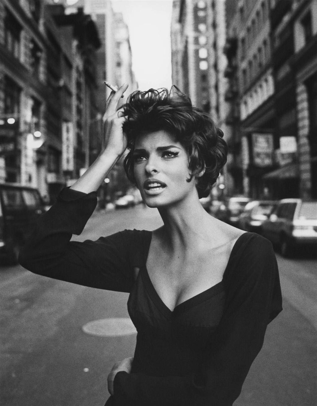 A woman in a black dress with a cigarette stands in the middle of a city street lined with high-rise buildings