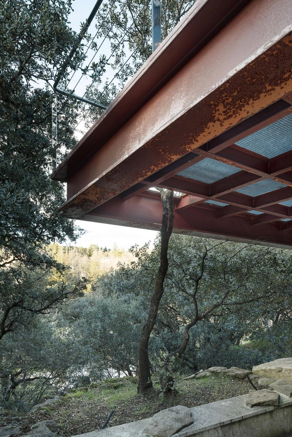 The underside of a weathered steel beam of a house hovering above the trees on a hillside