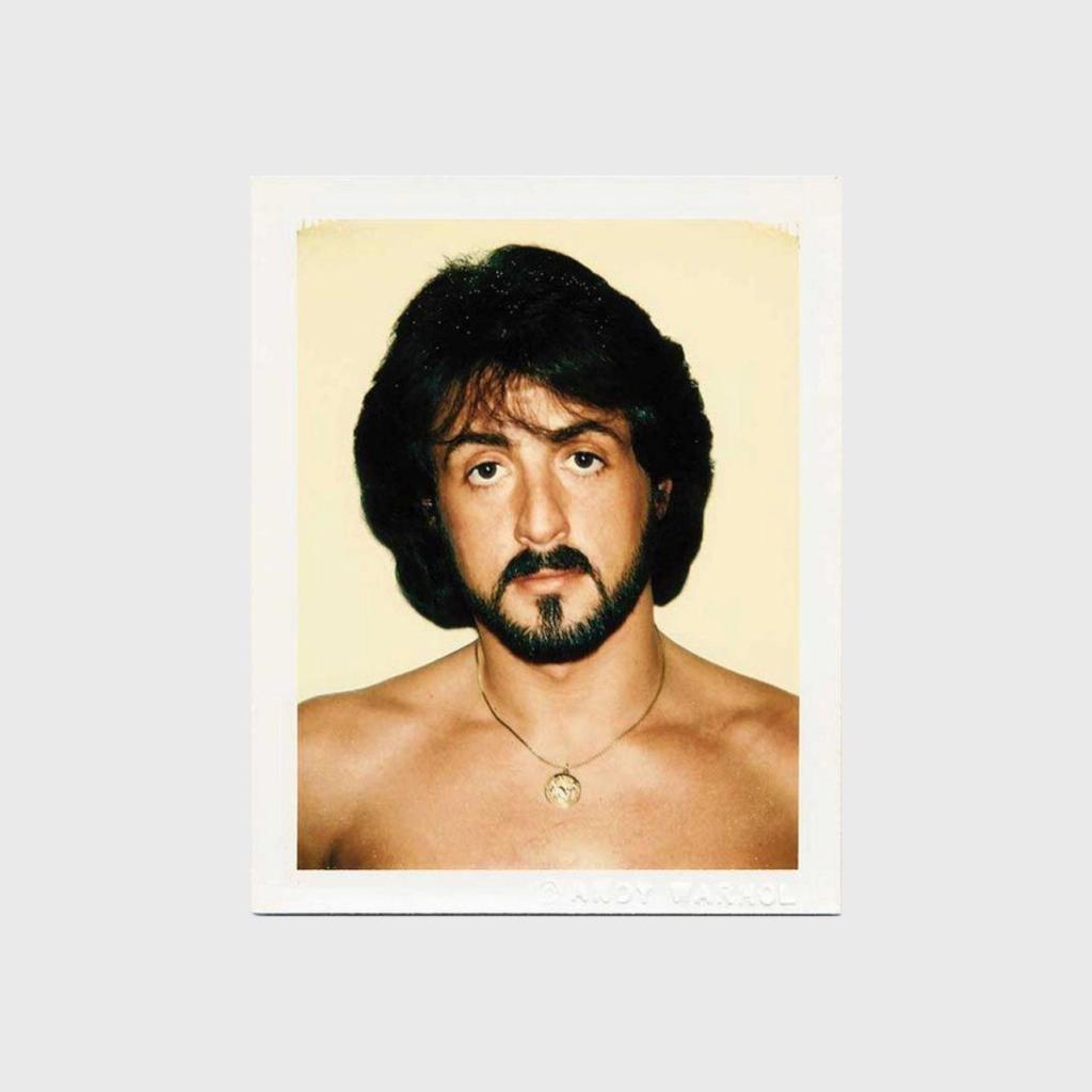 A Polaroid portrait of a bare-chested man with a goatee wearing a medallion on a chain