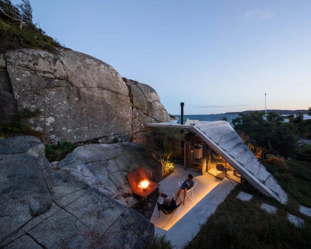 A coastal cabin with a pitched roof among granite rocks by the sea, a bonfire and people at dusk