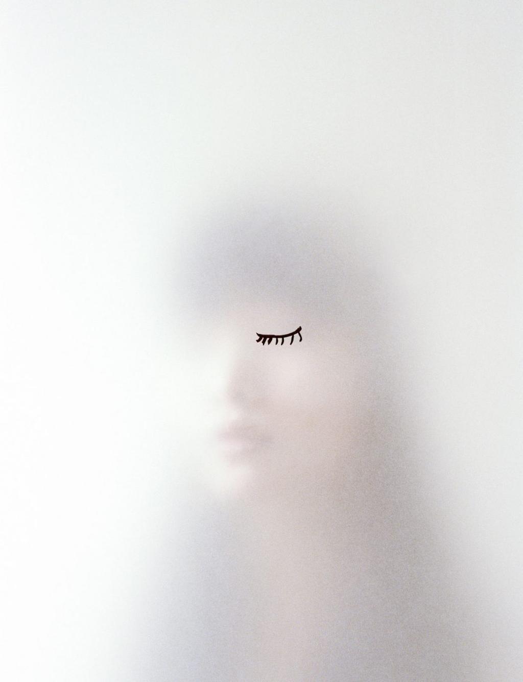 A blurred female face with one closed eye drawn in ink, against a white background