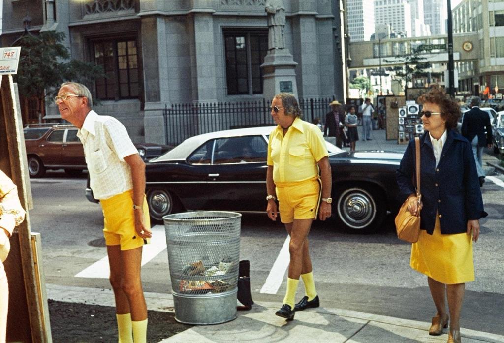 Colour street photograph: two men in yellow shorts and a woman beside a rubbish bin on a city corner