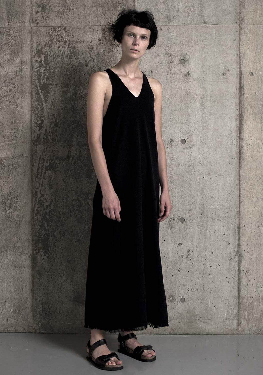 A model by a concrete wall in a long black sleeveless dress and sandals