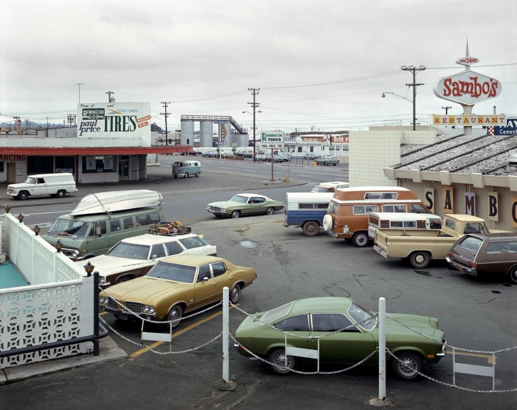 A car park filled with American cars from the seventies, in front of a Sambo's restaurant and a tyre shop