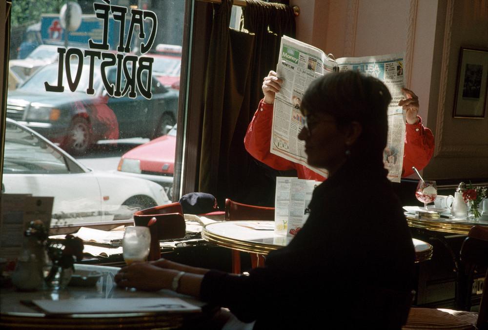 A woman in red reads a newspaper at Cafe Bristol, cars visible through the window