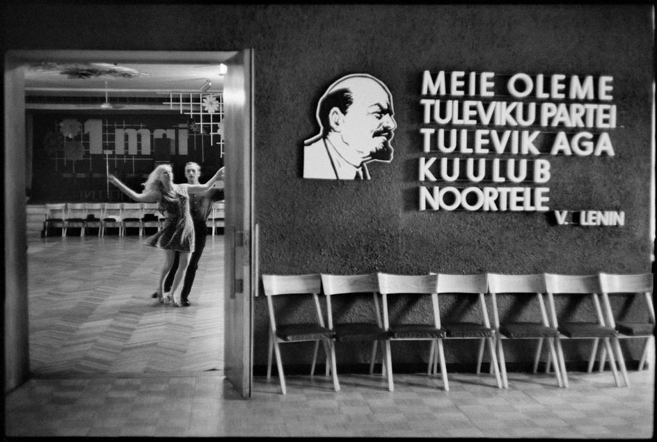 A couple dancing in a doorway beside a wall bearing a portrait of Lenin and one of his quotations