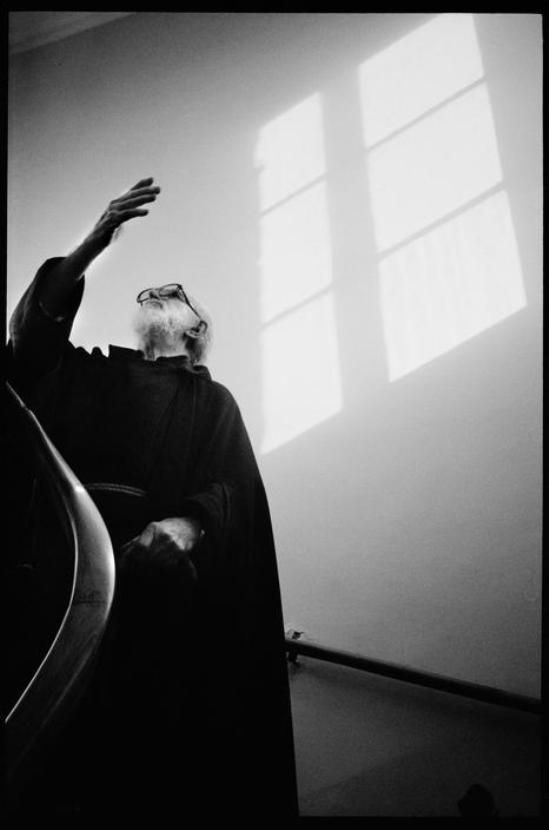 An elderly man in glasses raises his hand towards the sunlight coming through a window on a staircase