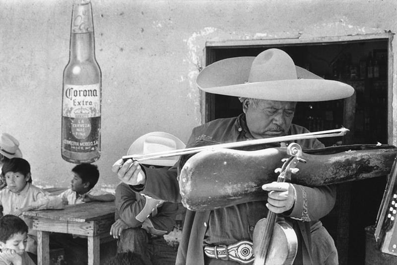 A musician in a sombrero plays violin against a wall bearing a Corona beer advertisement
