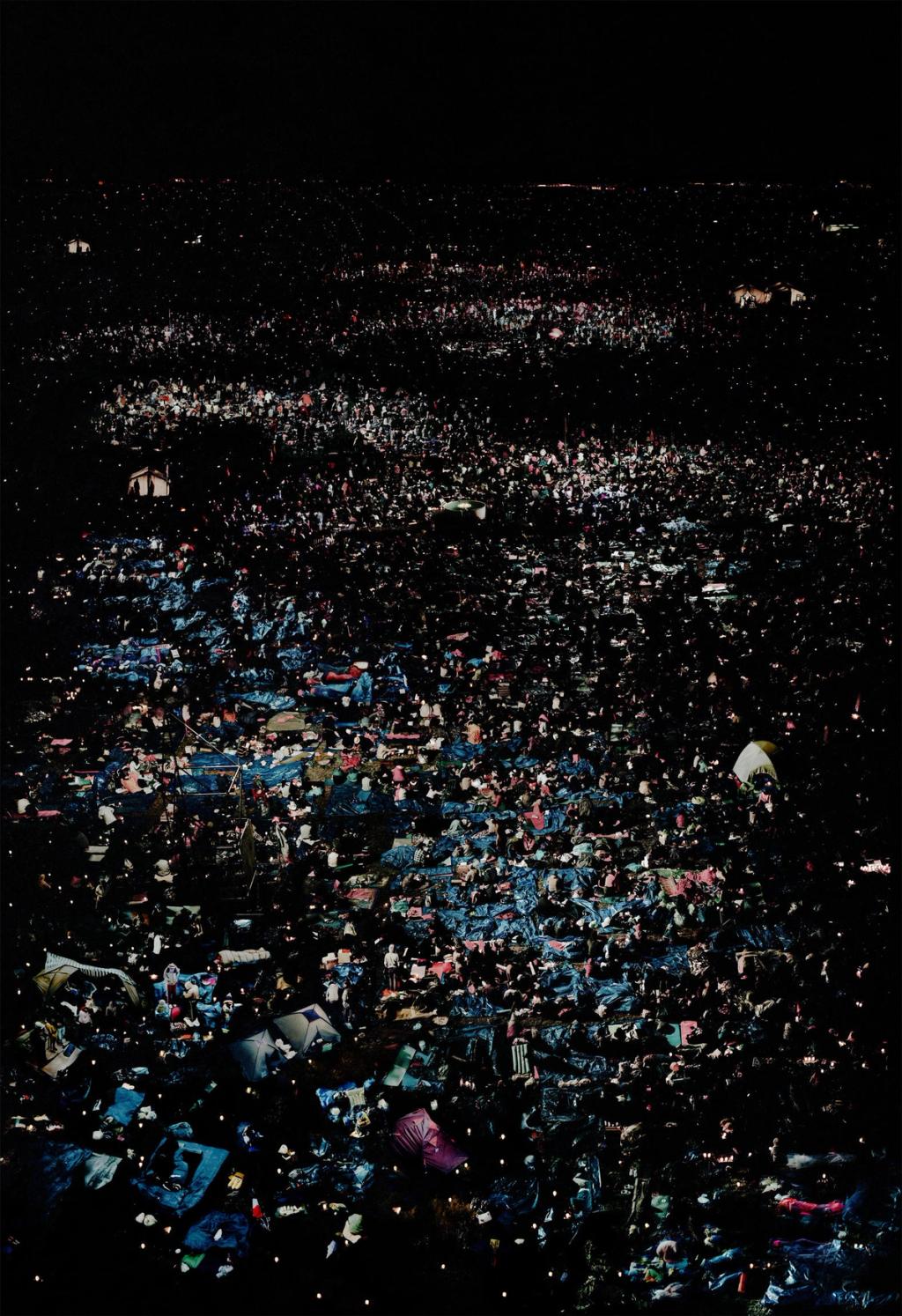 An open-air night concert from above: a dark field scattered with thousands of people and tents