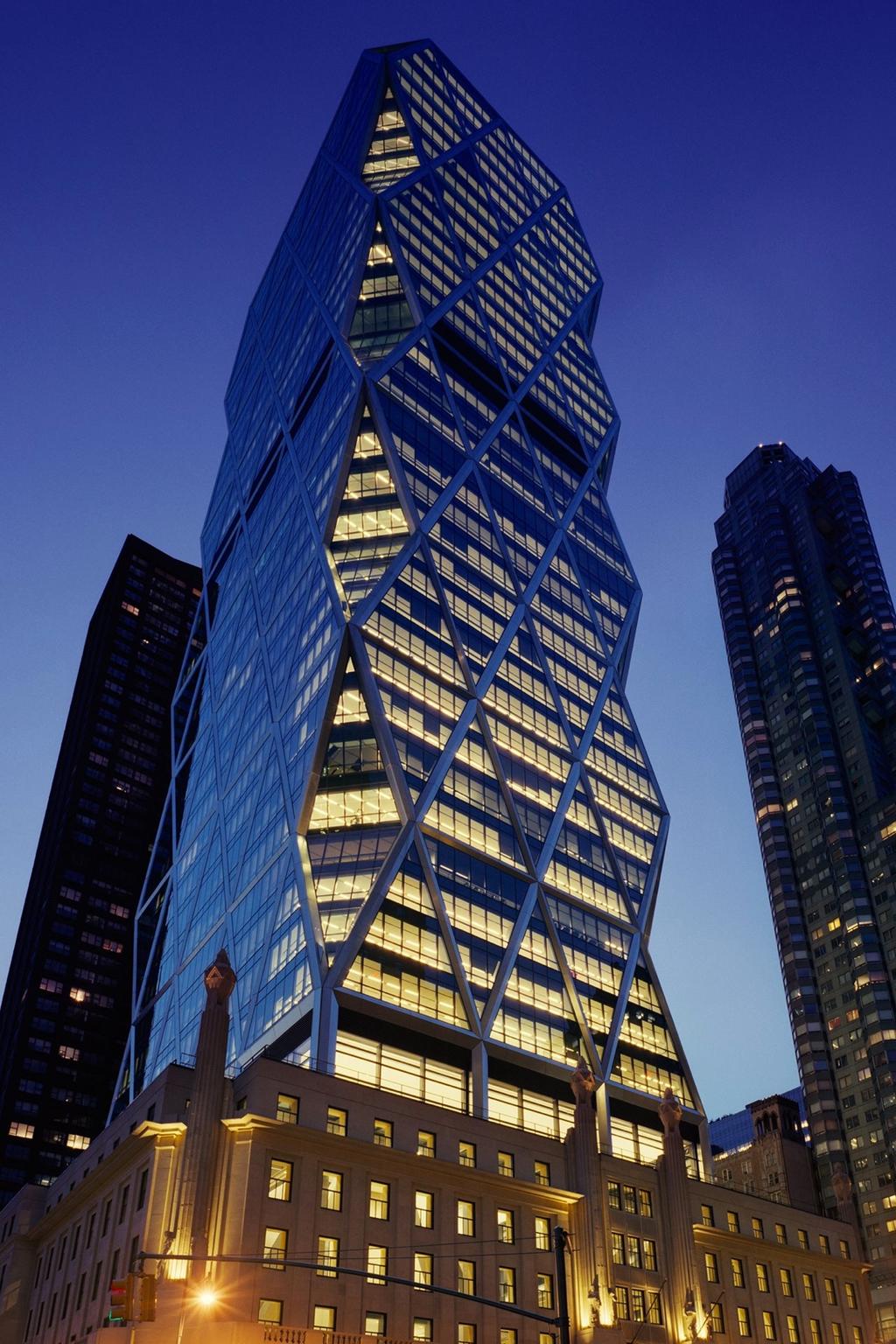 The Hearst Tower with its diagonal exoskeleton, lit up in the evening above its historic base