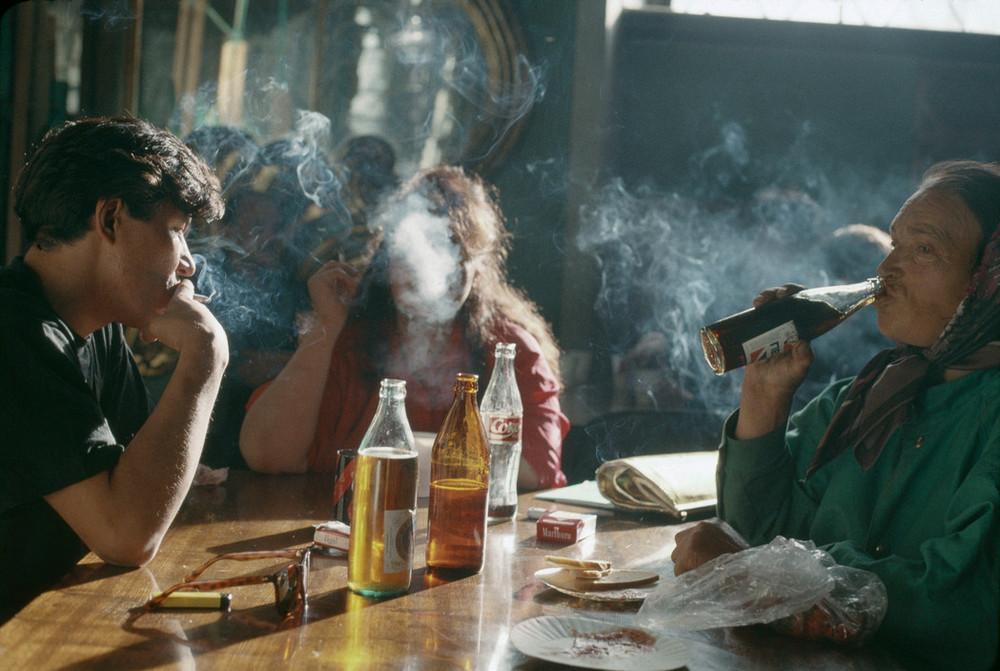 Three people at a table in a haze of cigarette smoke, an elderly woman drinking from a bottle