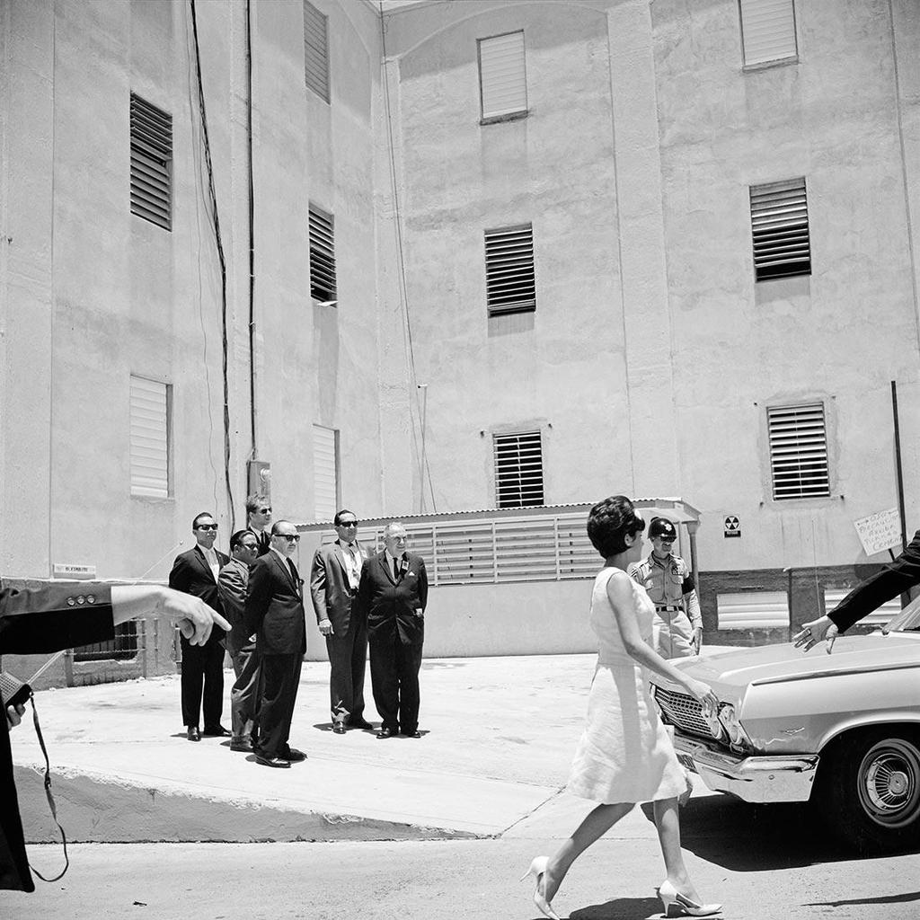 Black-and-white photograph: a group of men in suits and a woman walking toward a car against a blank wall