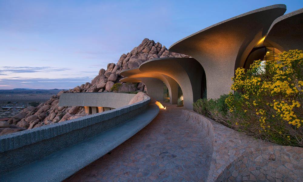 A curved pathway along the façade of a desert house with lighting and rock outcroppings at dusk