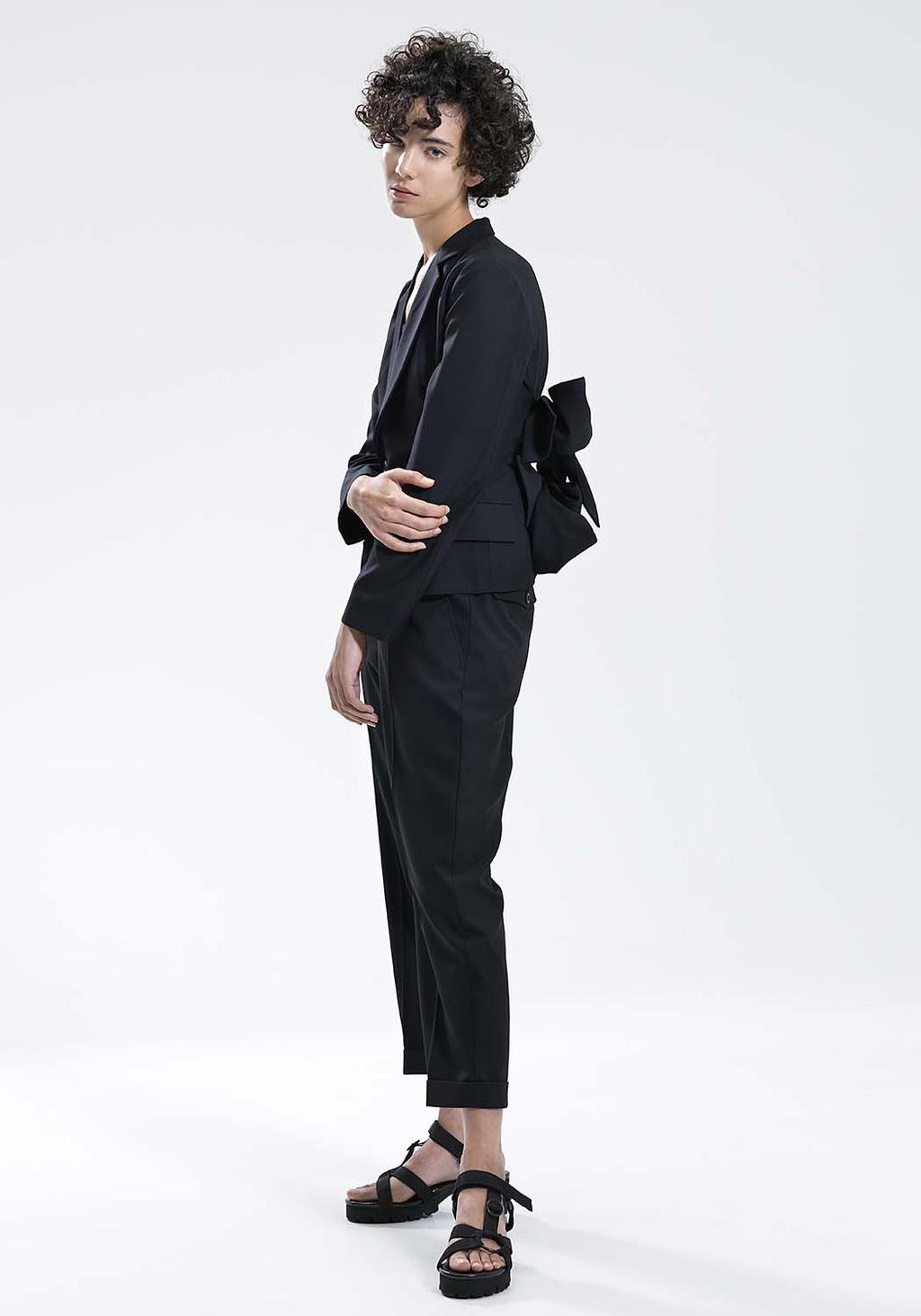 A model in profile in a black jacket with a bow at the back and tapered trousers