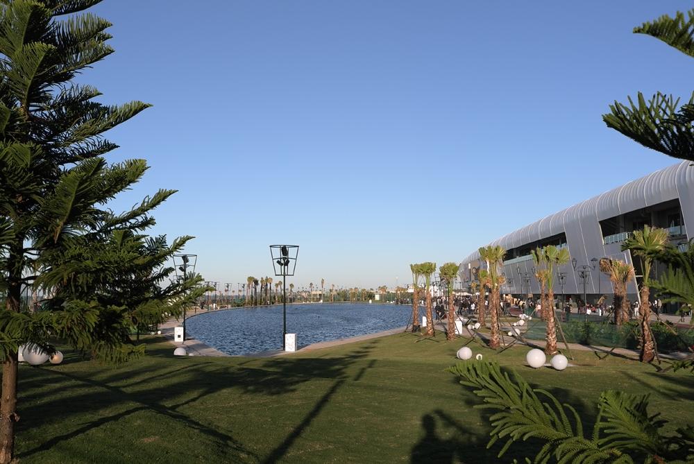 A lawn with palm trees and a pool in front of the curved Morocco Mall building during the day