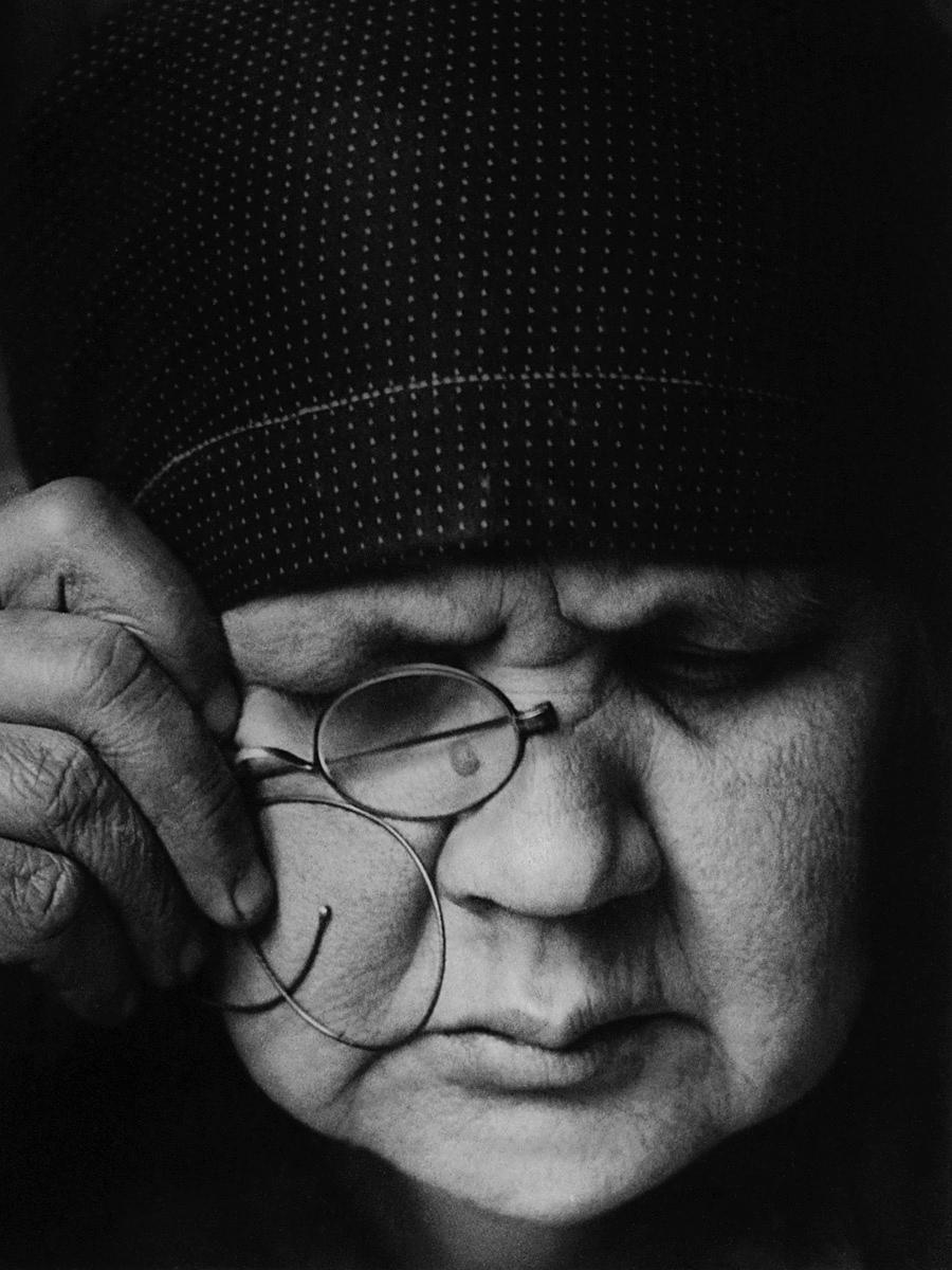 A portrait of a stern woman in a headscarf, one hand holding her glasses to her face