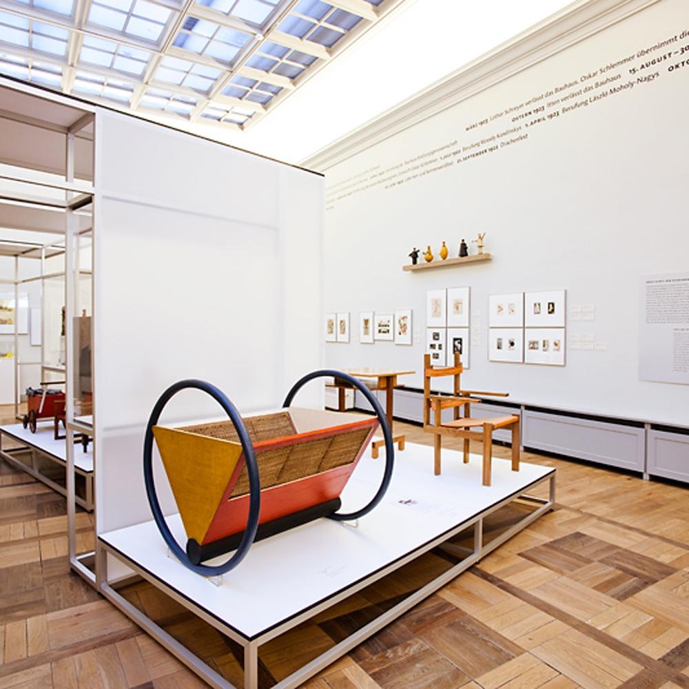 The geometric Bauhaus cradle on a museum plinth, surrounded by other furniture and drawings on the wall