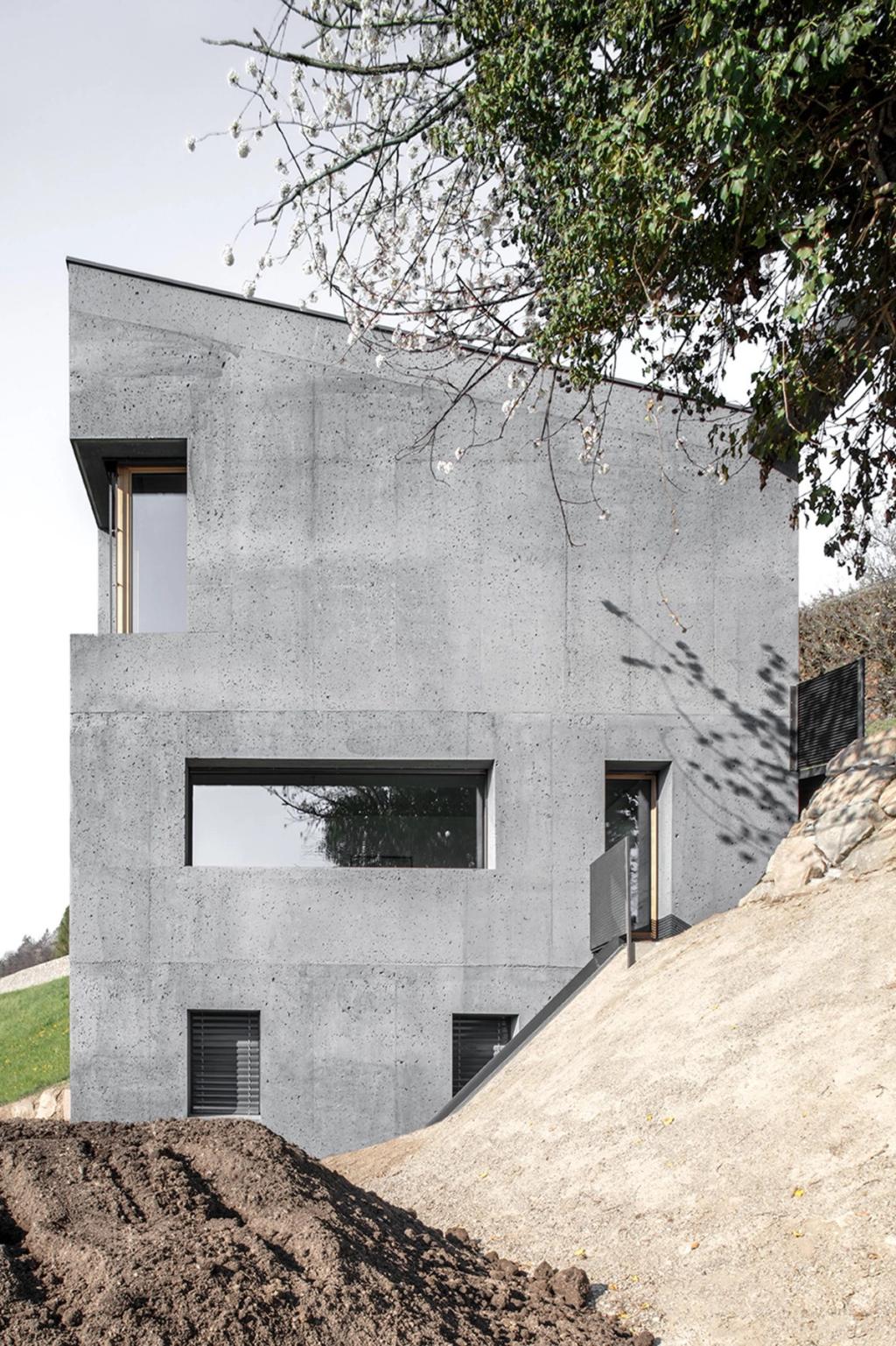 Grey concrete facade of the Habitat Andergassen Urthaler house with a horizontal window above an embankment