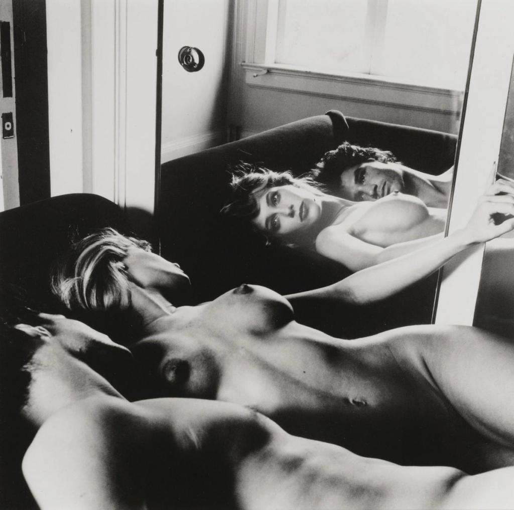 Two nude women lie side by side, reflected in a room mirror