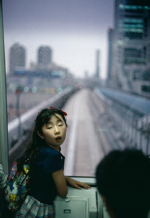 A girl with ribbons in her hair in a train carriage, the blurred skyscrapers of Tokyo beyond the window
