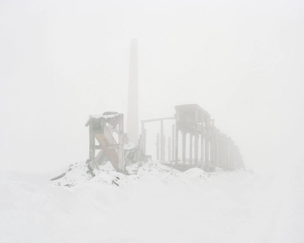 The ruins of a wooden industrial building with a chimney stack, shrouded in thick fog and snow