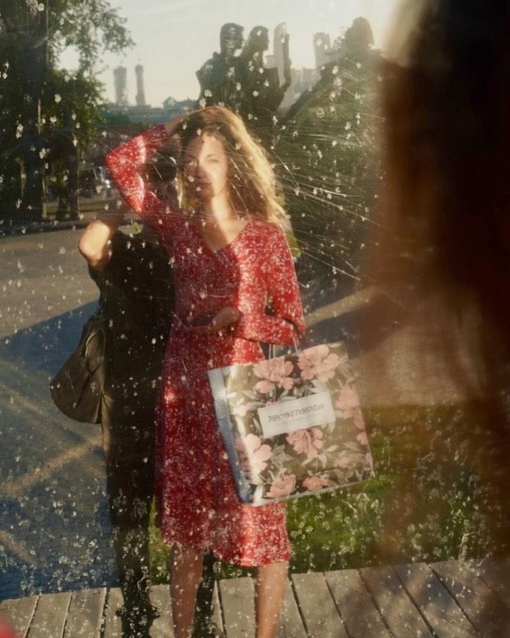 A woman in a red dress carrying a bag, seen through glass spattered with water drops at sunset