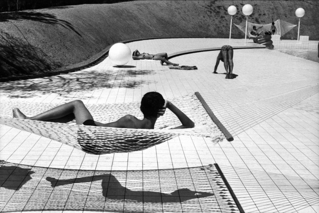 A woman in a hammock by a swimming pool, surrounded by a tiled terrace and white globe lamps