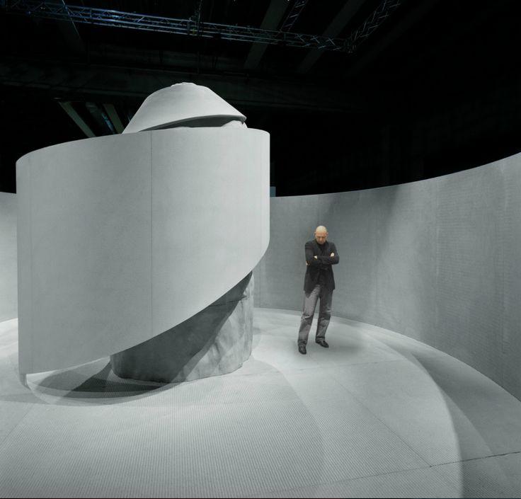 A man in a dark suit standing beside a white cylindrical installation in an exhibition hall