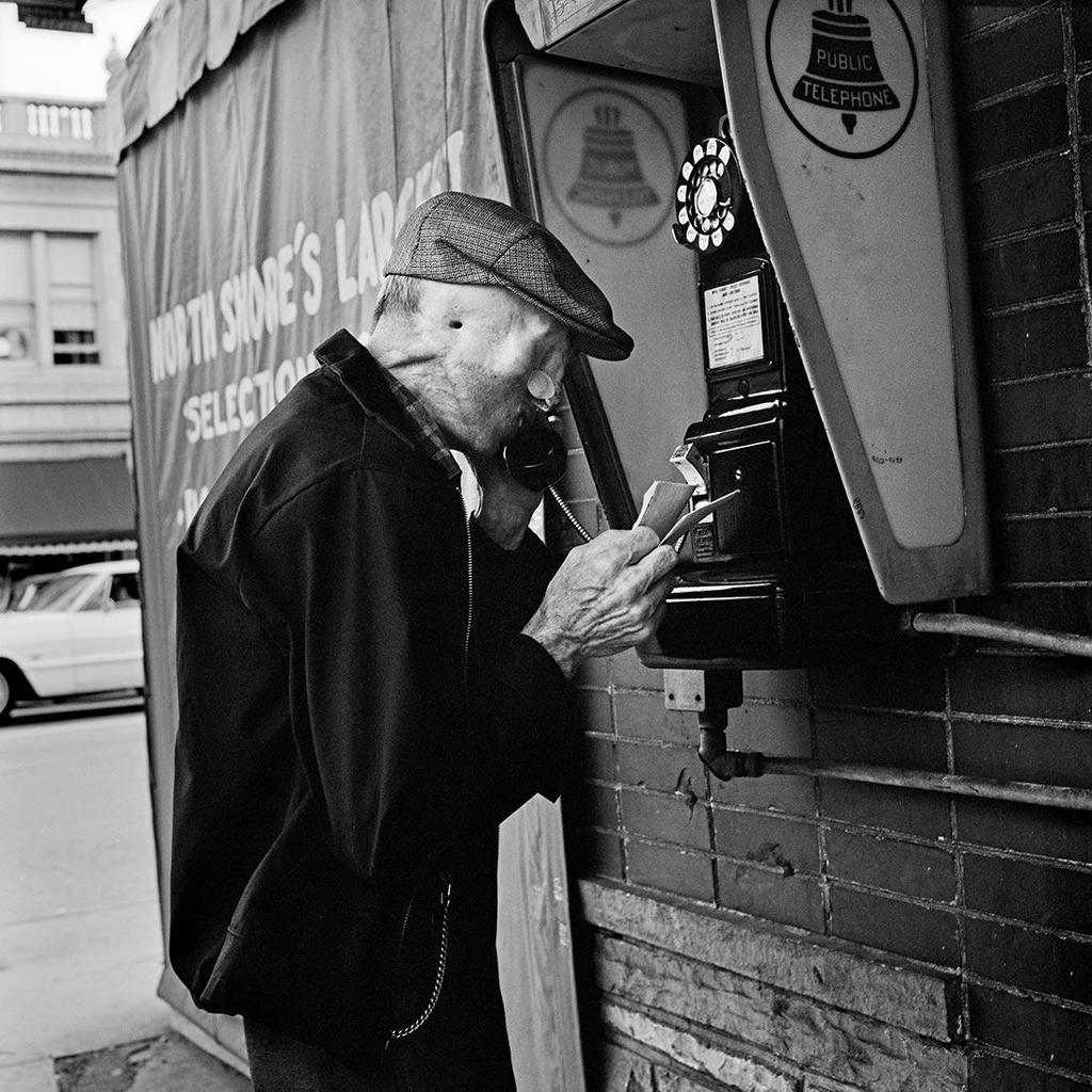 Black-and-white photograph: an elderly man in a cap talks on a street payphone beside a brick wall