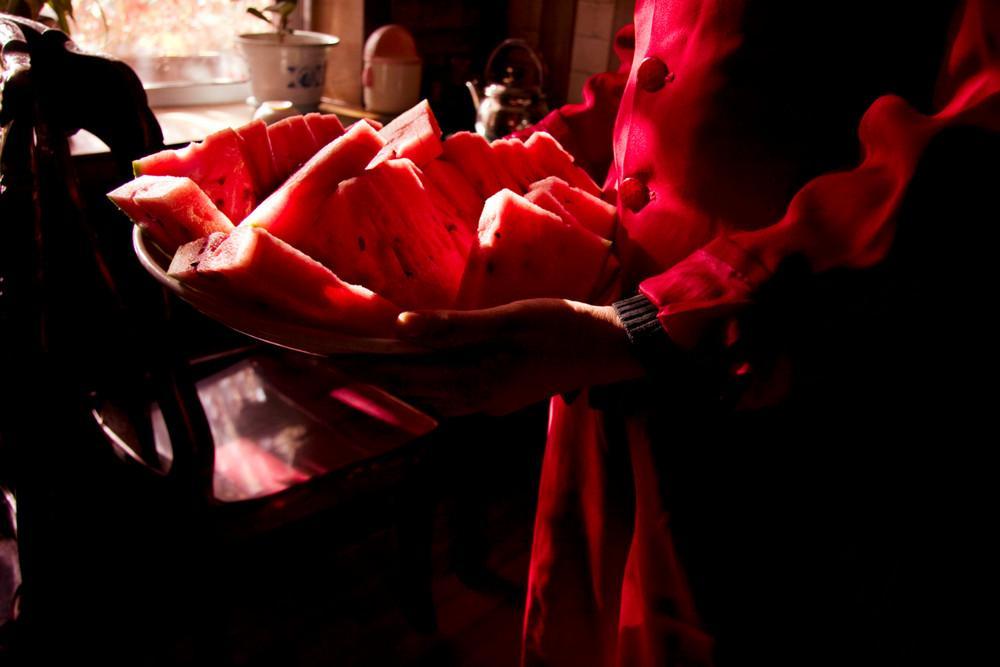 Hands holding a dish of watermelon slices in a dark interior, reddish light