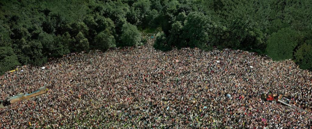 A vast crowd gathered in a clearing surrounded by dense green forest, seen from above
