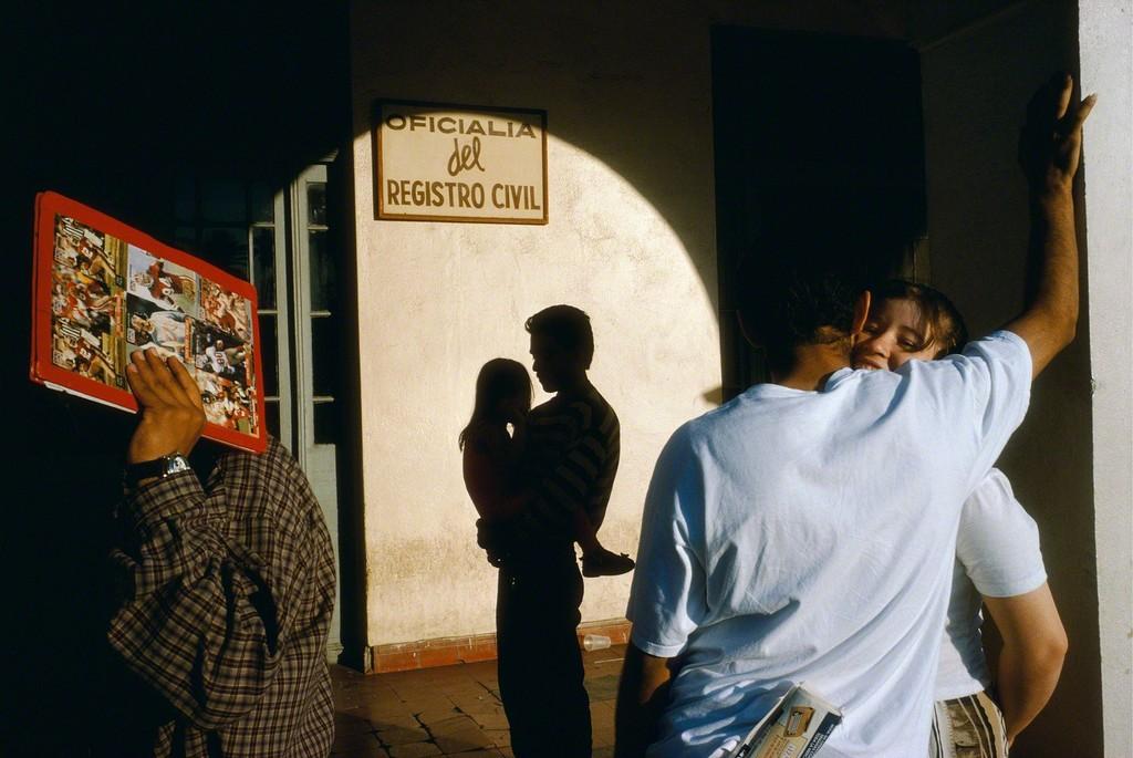 A sun-drenched street at an entrance bearing the sign Registro Civil, a couple embracing and the silhouette of a person with a child