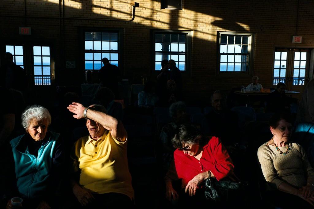 Elderly people sit in a hall cut through by bright strips of light from windows overlooking the sea