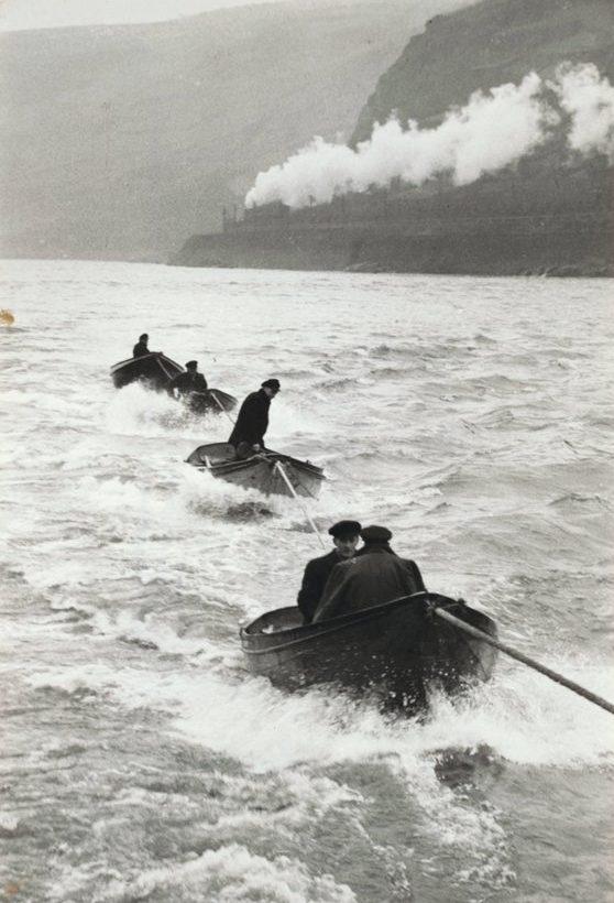 Men in rowing boats on choppy water, a steam locomotive passing along the bank