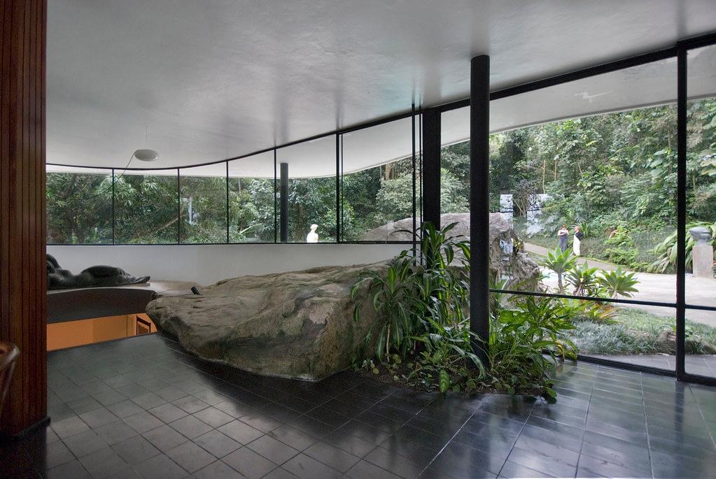 The living room of Casa Canoas with a granite boulder rising inside and panoramic glazing opening onto the forest