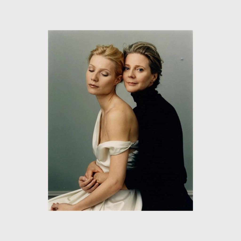 A colour portrait of two women standing back to back, one in a light dress and the other in a black turtleneck