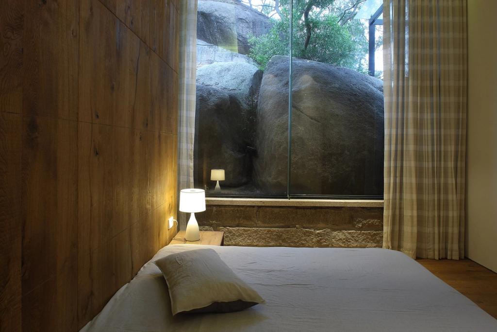 A bedroom with a wooden wall and a panoramic window looking onto boulders and trees beyond the glass