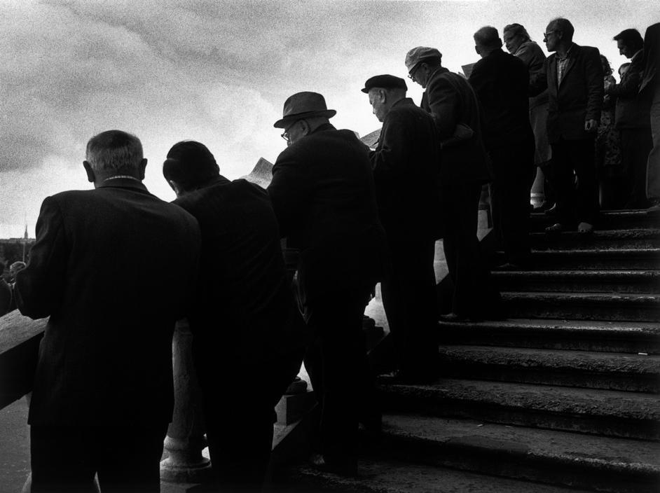 A group of elderly men in overcoats climbing stone steps against the sky
