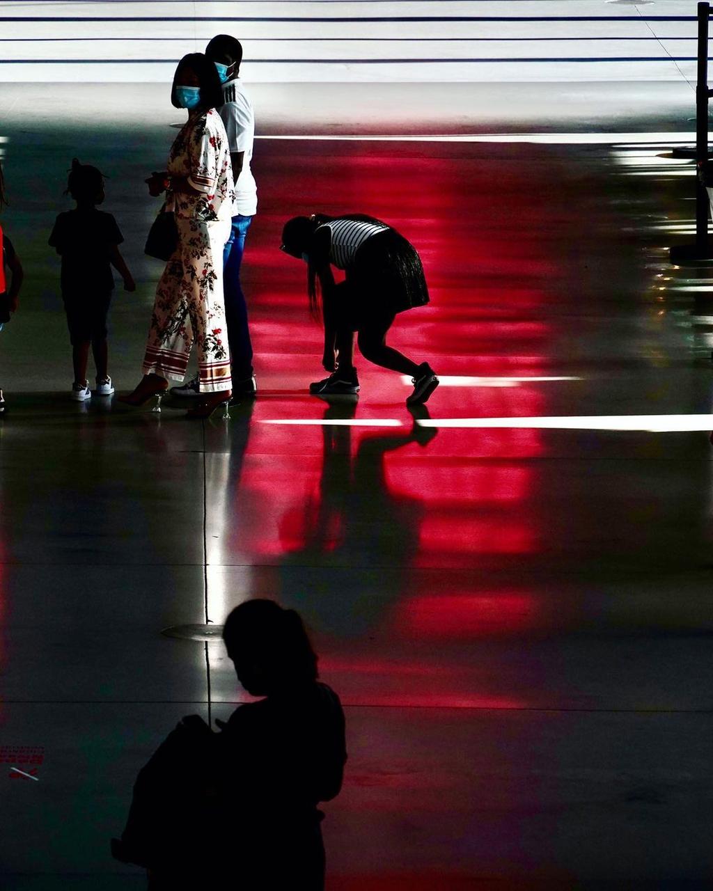 Silhouettes of people in a hall, a stooping figure caught in a band of red light on the floor