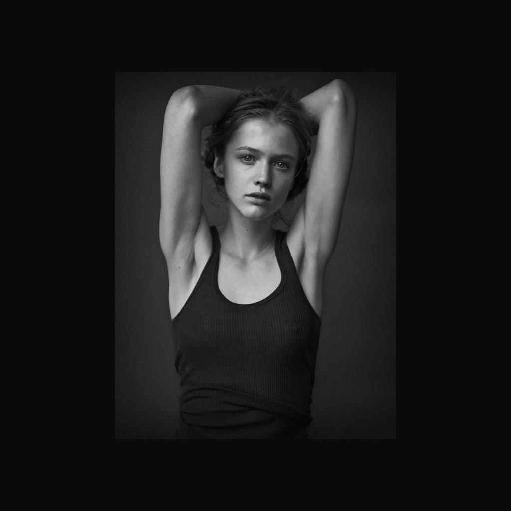 A black-and-white portrait of a young woman in a black vest top with her arms raised behind her head