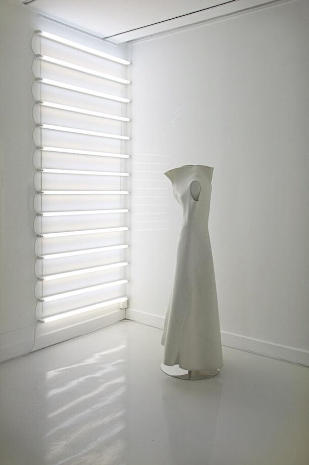A white sculptural dress displayed against a wall of fluorescent lights in a gallery
