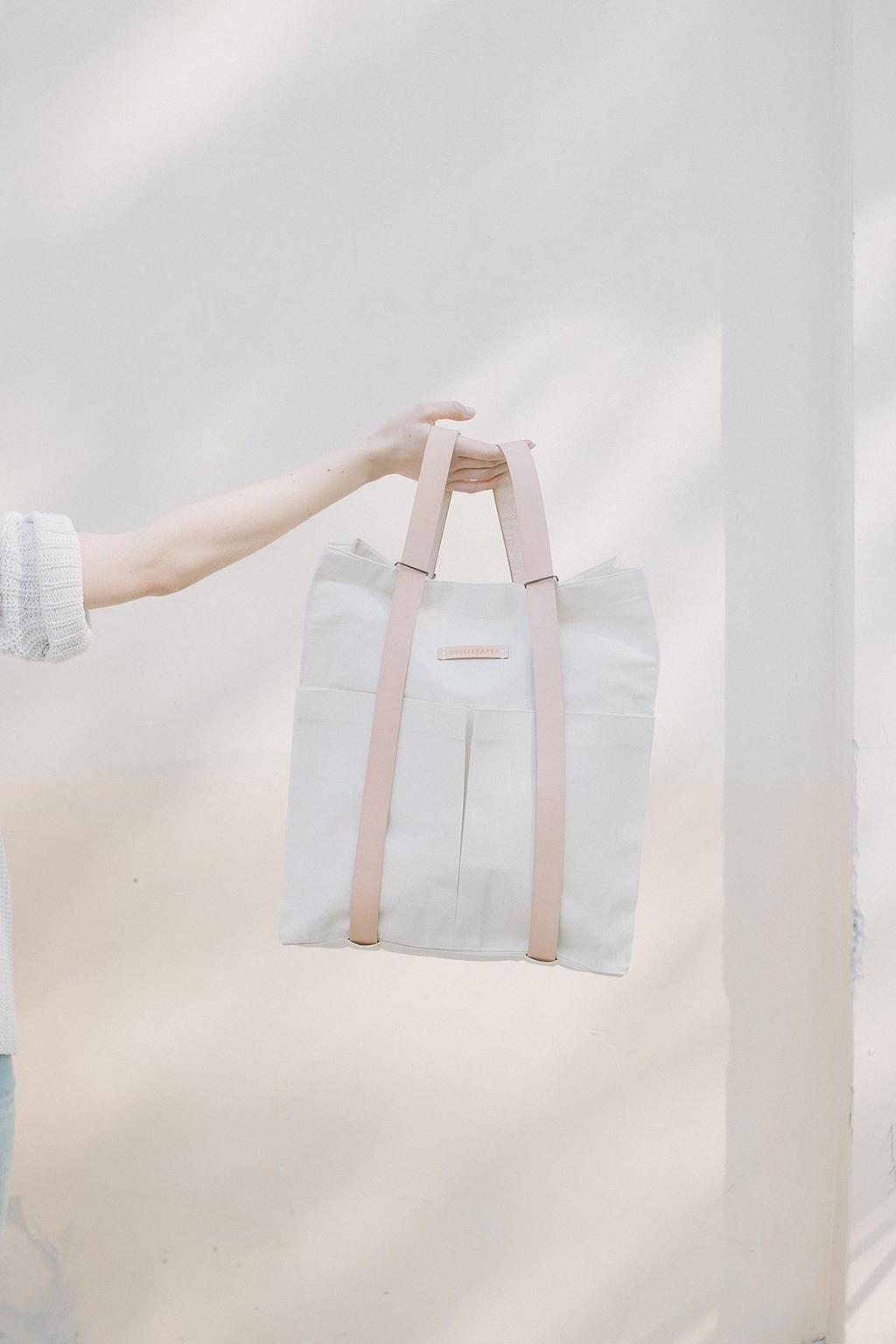 A light canvas Thisispaper tote bag with leather handles held in an outstretched hand