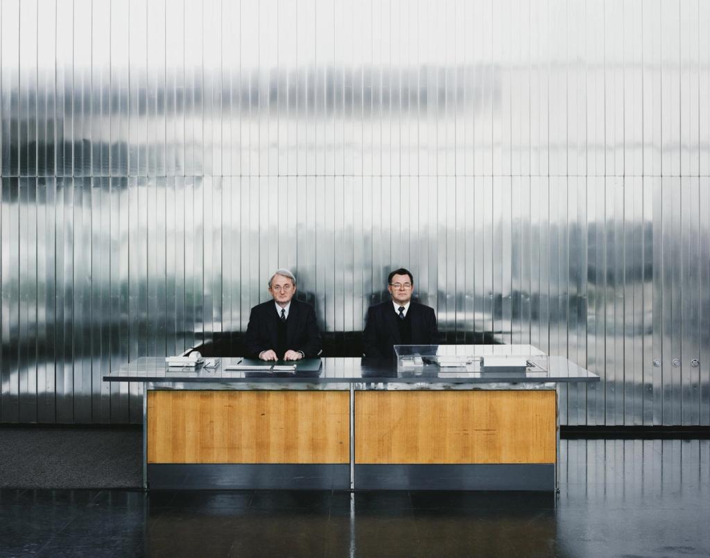 Two men in dark suits seated at a table against a wall of frosted ribbed glass