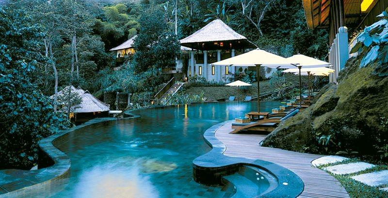 Изогнутый бассейн отеля Ubud Hanging Gardens с деревянным настилом среди тропической зелени
