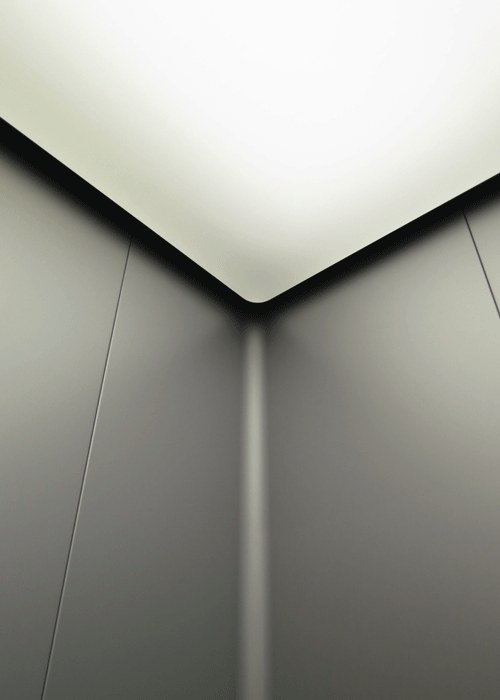 Corner of a lift cabin with grey panels and an illuminated ceiling
